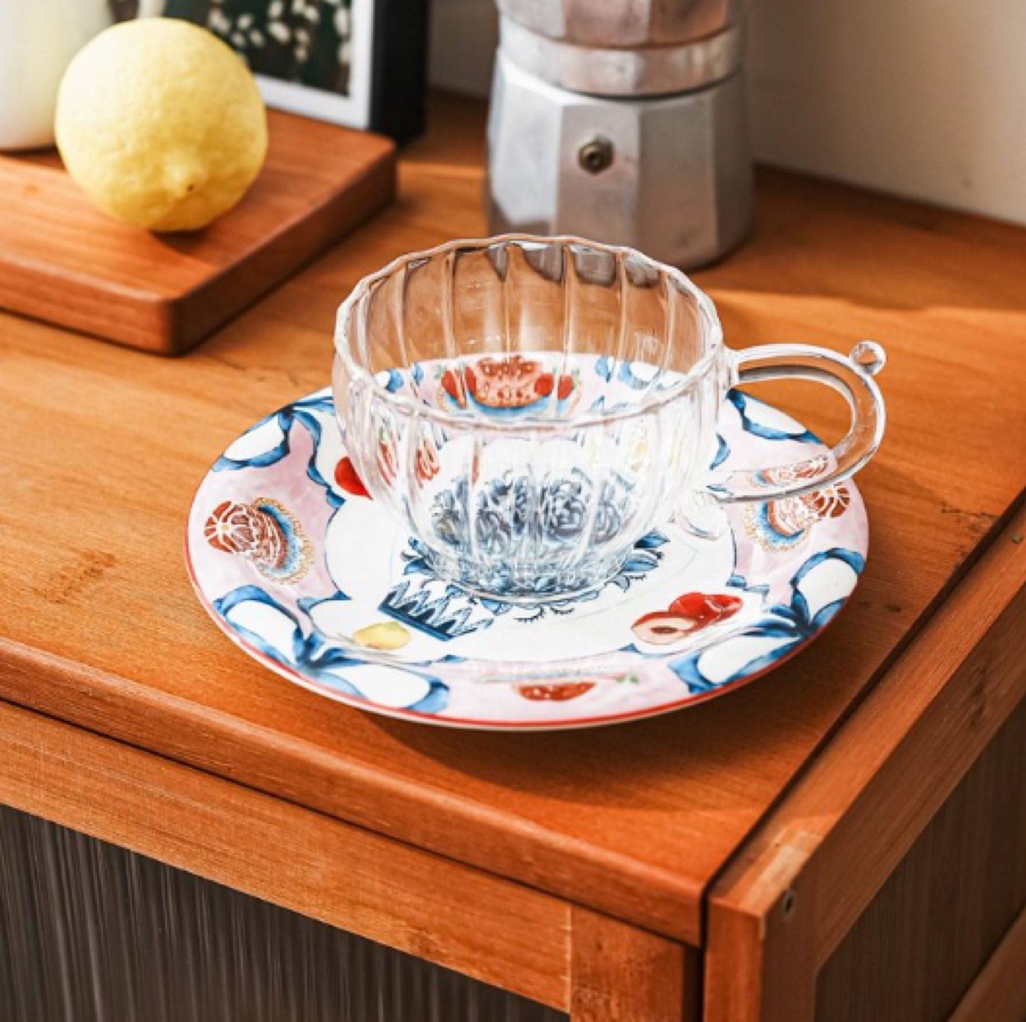 Glass cup and ceramic saucer on a kitchen counter beside a lemon and a coffee grinder.